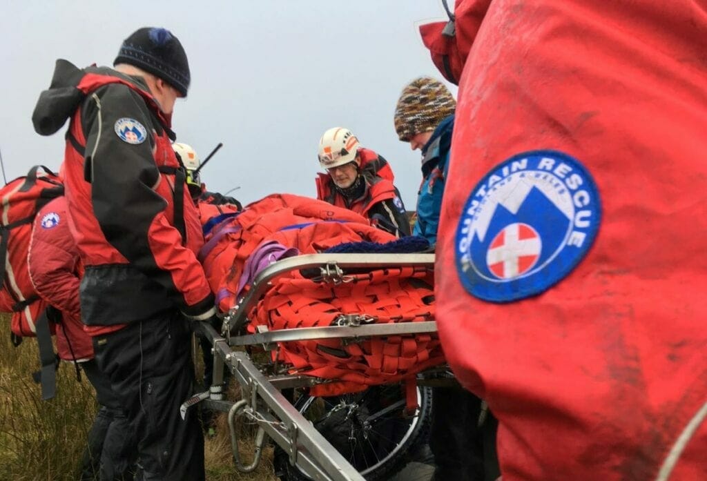 Penrith Mountain Rescue Team carry the woman on a stretcher. The sky is grey and the rescuers are wearing the red and black mountain rescue jackets